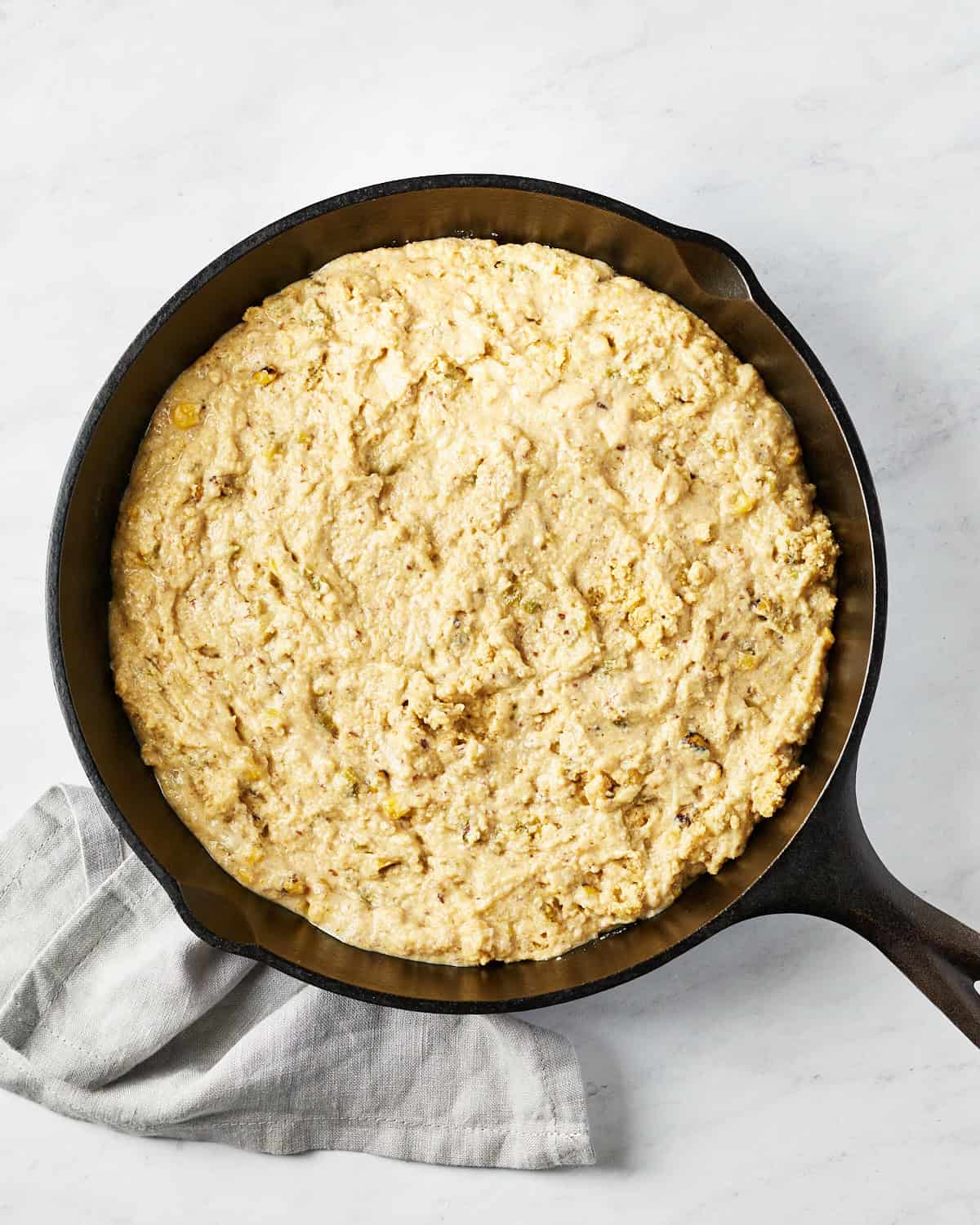 Overhead view of cornbread batter in cast iron skillet on marble background and grey napkin