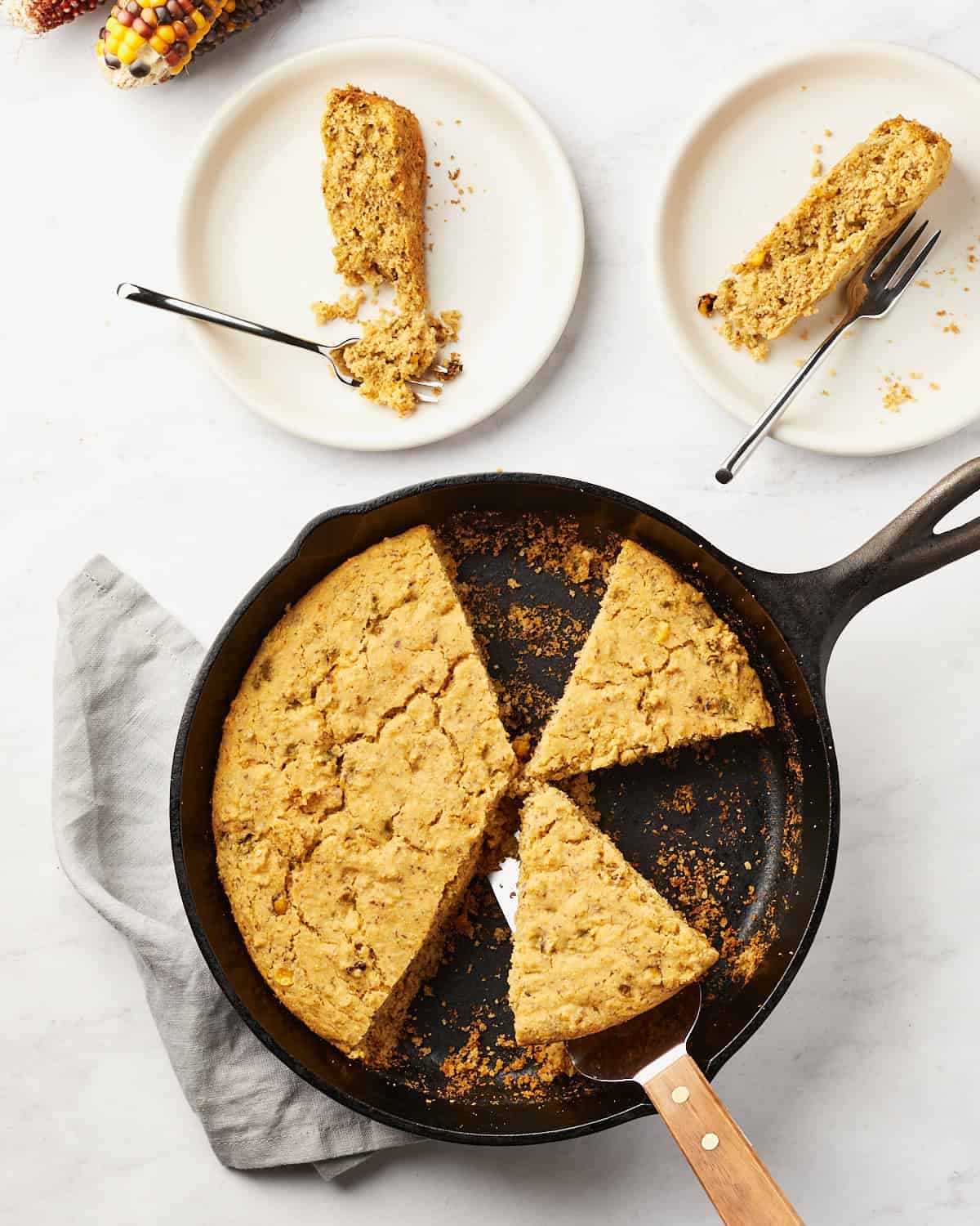 Baked Vegan cornbread in skillet and on two pieces on small plates on marble background.