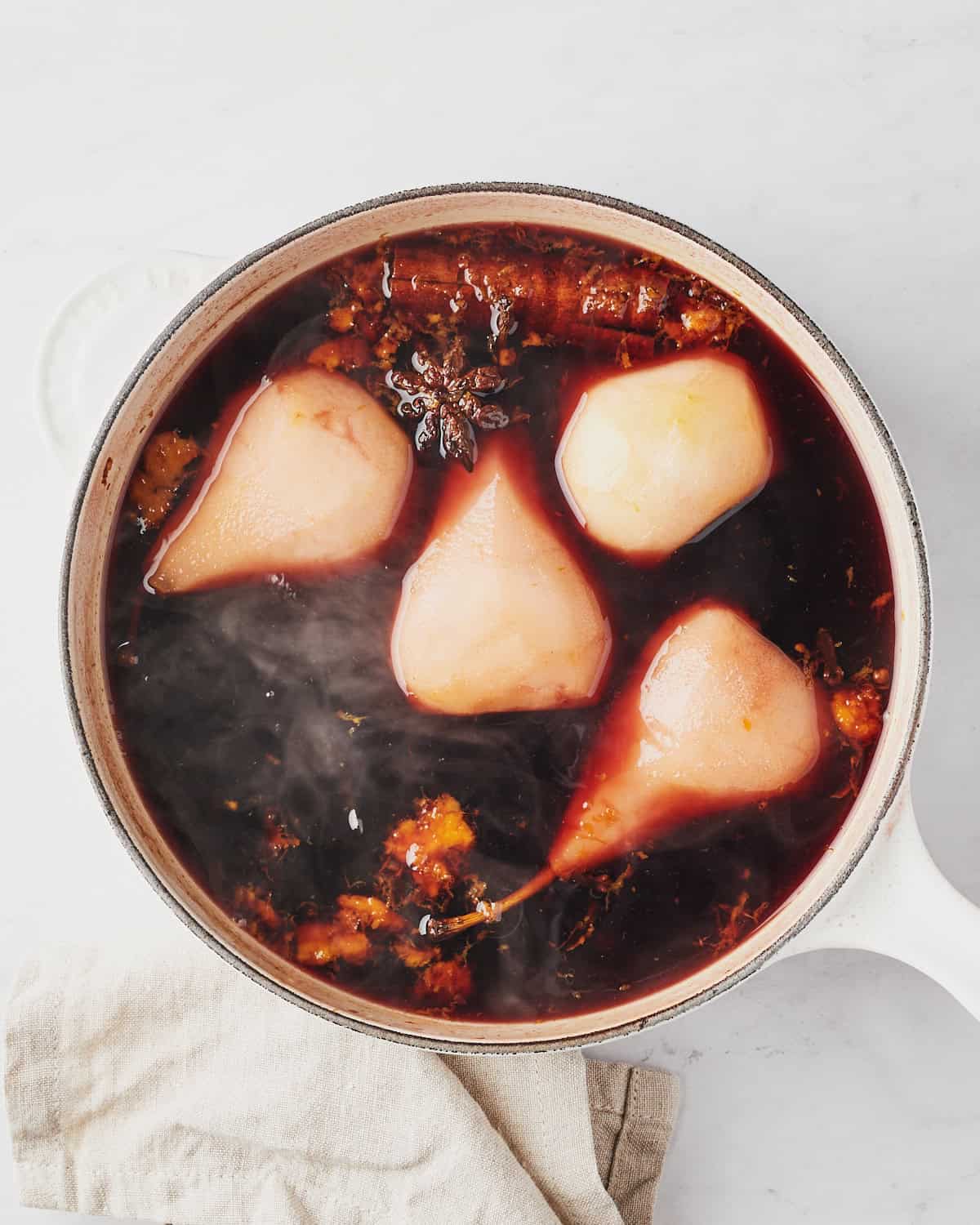 Four peeled pears placed in a red wine broth with spices in a saucepan.