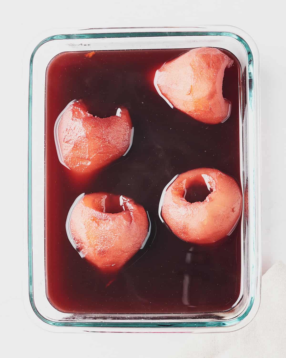 Four red wine poached pears placed in a glass container with red wine poaching liquid.