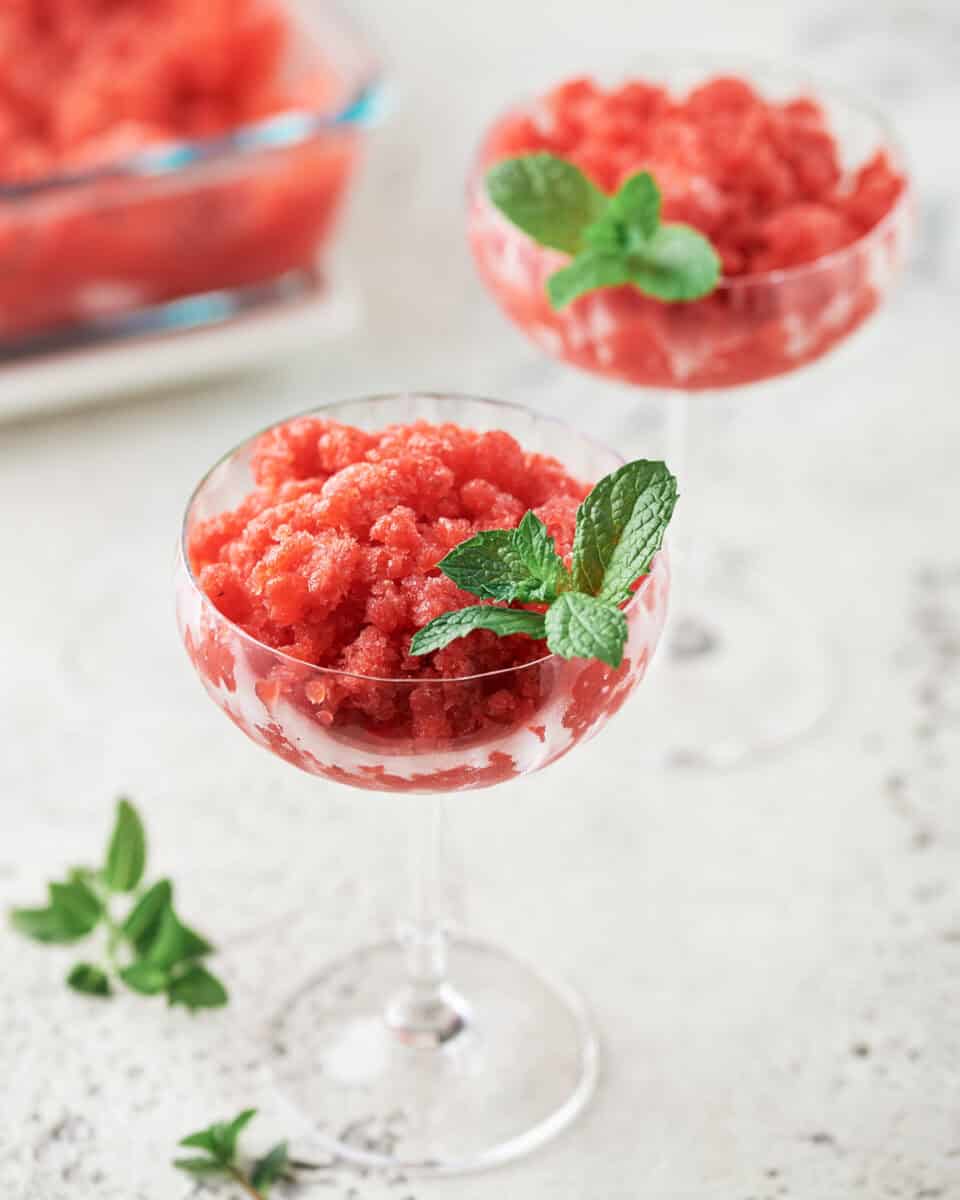 Two coupe glasses filled with strawberry lemon granita with gin accented with sprig of mint.