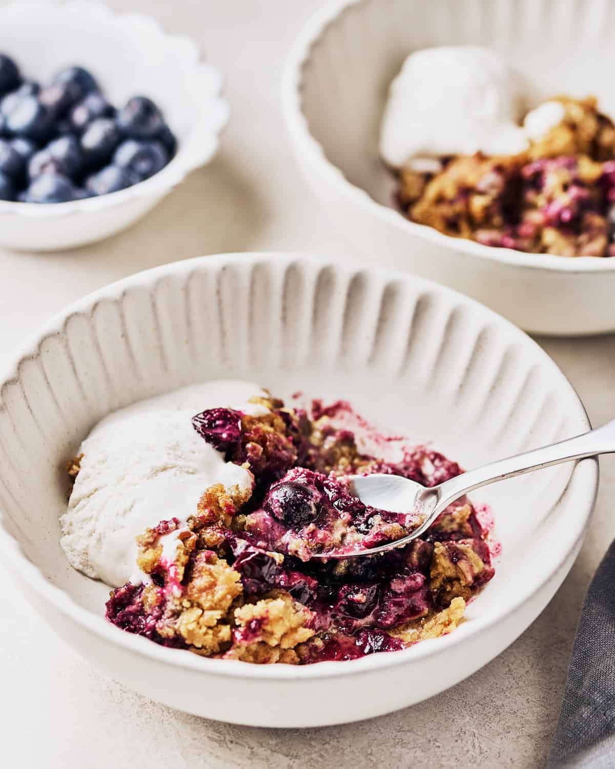 Two bowls of vegan blueberry cobbler, one close up with a spoon in it with a side of vegan ice cream.