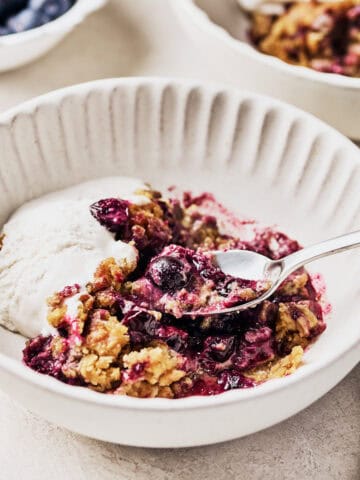 A bowl of vegan blueberry cobbler, close up with a spoon in it with a side of vegan ice cream.