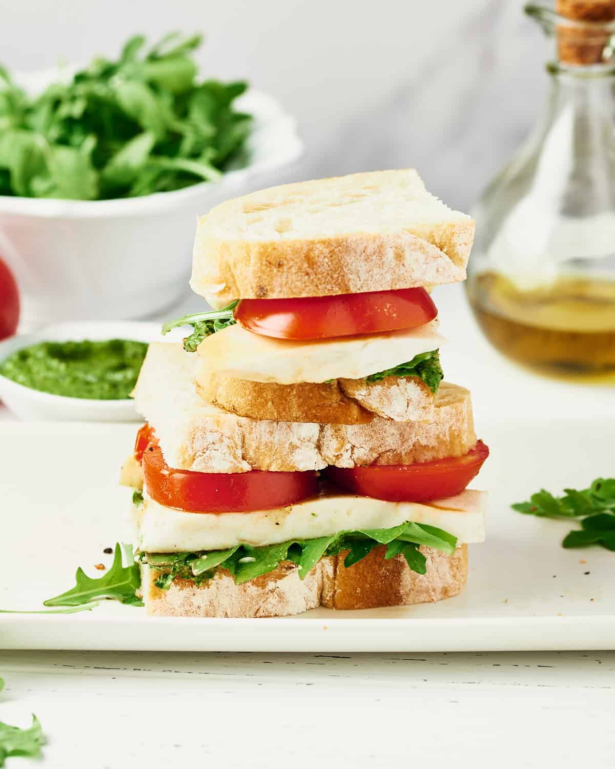Stacked Vegan Caprese Sandwich with sliced tomatoes, arugula, vegan pesto, and vegan mozzarella.