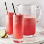 Two glasses of watermelon water next to a pitcher on a round platter with half a lime.