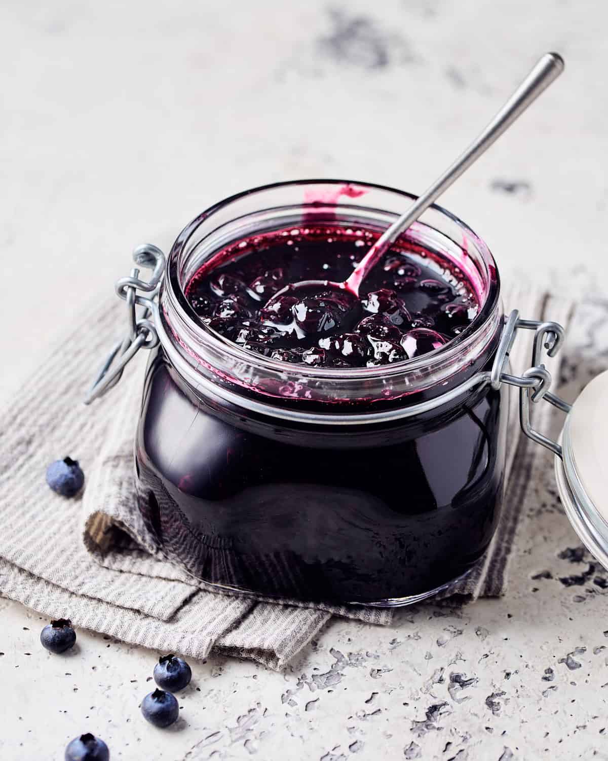 Blueberry Compote in a glass jar with a spoon in it on a napkin with blueberries nearby.