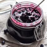 Blueberry Compote in a glass jar with a spoon in it on a napkin with blueberries nearby.