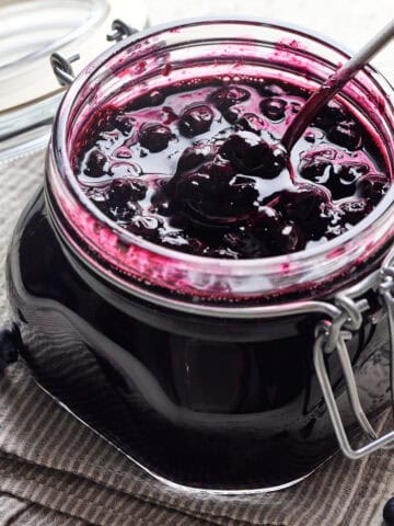 Blueberry Compote in a glass jar with a spoon in it on a napkin with blueberries nearby.