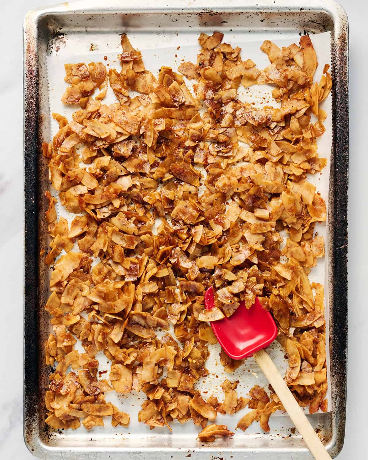 Stirring coconut bacon with a spatula on a rimmed baking sheet.